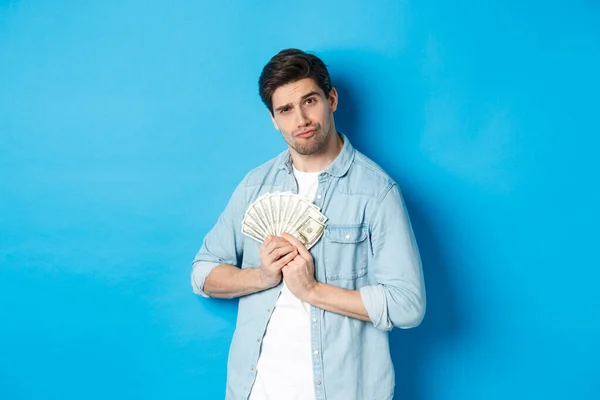 Joven guapo guardando dinero para sí mismo, sonriendo y luciendo codicioso, de pie sobre fondo azul — Foto de Stock