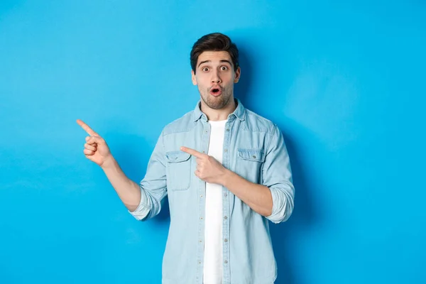 Retrato del hombre adulto sorprendido con ropa casual mostrando el anuncio, señalando con los dedos hacia la izquierda y mirando asombrado, de pie sobre fondo azul — Foto de Stock