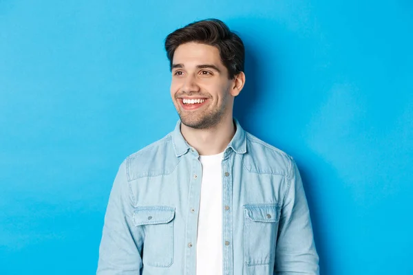 Young modern guy with beard, looking left and smiling pleased at copy space, standing over blue background