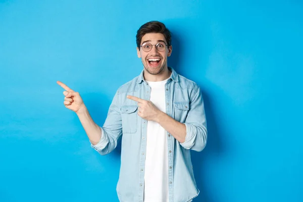 Hombre guapo excitado en gafas apuntando con los dedos hacia la izquierda, mirando feliz mientras muestra publicidad, demostrando espacio de copia sobre fondo azul — Foto de Stock