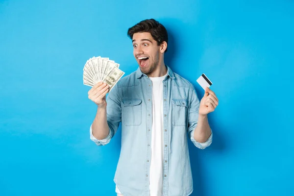Hombre sorprendido y feliz con tarjeta de crédito, mirando el dinero satisfecho, de pie sobre fondo azul — Foto de Stock