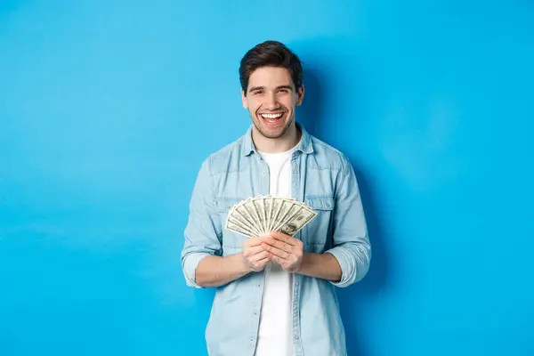 Feliz hombre exitoso sonriendo complacido, sosteniendo el dinero, de pie sobre fondo azul — Foto de Stock