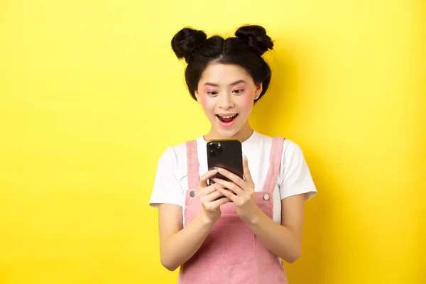 Bella ragazza asiatica guardando eccitato allo schermo, la lettura di messaggi al telefono e sorridente felice, in piedi in abiti estivi su sfondo giallo — Foto Stock