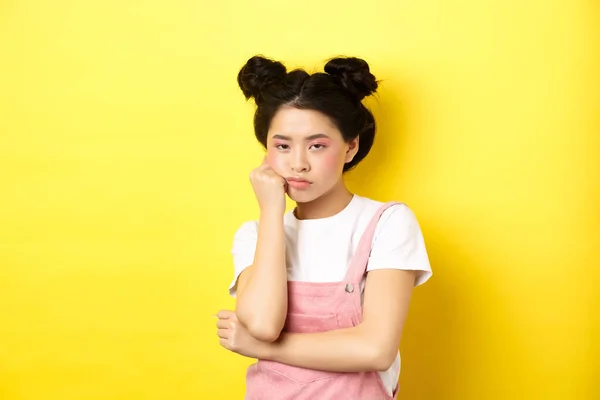 Скучная азиатская девушка-подросток со стильным макияжем и летней одеждой, выглядящая неохотно и равнодушной, стоящая на желтом фоне — стоковое фото