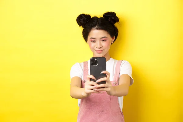 Elegante chica asiática tomando fotos en el teléfono inteligente, haciendo video con el teléfono celular y sonriendo, de pie sobre fondo amarillo — Foto de Stock