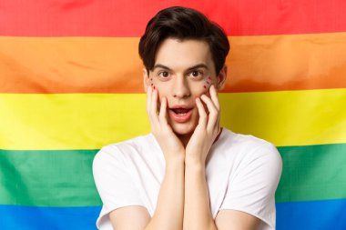 Beyaz tişörtlü, parlak makyajlı, kameraya bakıp eğlenen, yüzüne dokunan, gökkuşağı gurur bayrağına karşı duran güzel esmer gay adamın yakın çekimi.