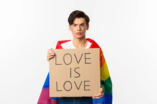 Foto cintura-up de joven activista masculino lgbtq, con bandera de arco iris y sosteniendo el amor es signo de la tarjeta de amor para el desfile del orgullo, la lucha por los derechos humanos, fondo blanco —  Fotos de Stock