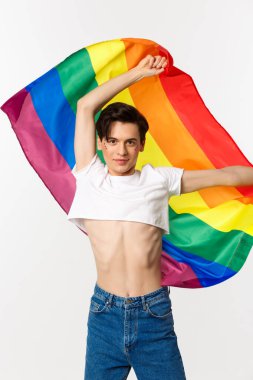 Ekin bluzlu ve kot pantolonlu mutlu eşcinsel kişinin dikey görüntüsü gökkuşağı bayrağını sallıyor, lgbtq bayramını kutluyor, beyaz arka planda duruyor.