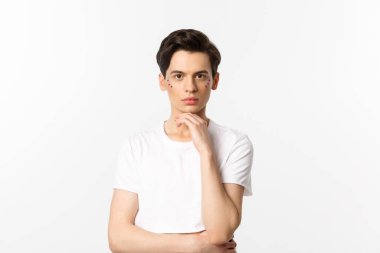 Beyaz tişörtlü, yüzünde sim olan ve kameraya ciddi ciddi bakan beyaz arka planda duran çekici bir gay resmi.