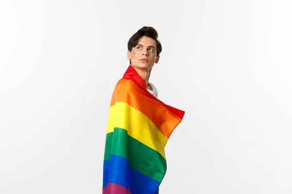Bild av omtänksam ung homosexuell man bär lgbtq flagga, vrida huvudet i övre vänstra hörnet, stirrar på logotypen, står över vit bakgrund — Stockfoto