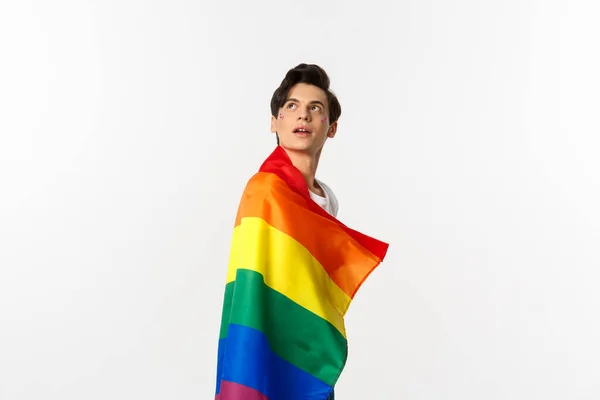 Derechos humanos y concepto de comunidad lgbtq. Hermoso joven andrógino con brillo en la cara, con bandera del orgullo del arco iris y mirando a la esquina superior izquierda, fondo blanco —  Fotos de Stock