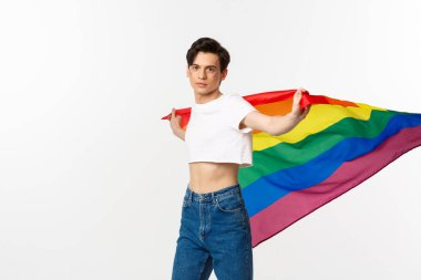 İnsan hakları ve lgbtq toplum kavramı. Gururlu bir eşcinsel gökkuşağı bayrağı sallıyor ve kameraya özgüvenli bakıyor. Kırpılmış bluz ve kot pantolon içinde beyaz arka plana karşı duruyor.