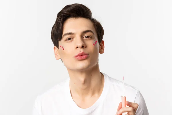 Close-up of beautiful gay man with glitter on face pucker lips after applying lip gloss, standing over white background — Stock Photo, Image