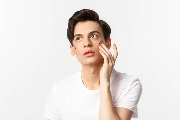 Άνθρωποι, κοινότητα Igbtq και την έννοια του τρόπου ζωής. Όμορφη νεαρός γκέι άντρας εφαρμογή glitter κάτω από τα μάτια για το κόμμα υπερηφάνεια, στέκεται πάνω από λευκό φόντο — Φωτογραφία Αρχείου