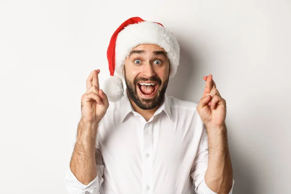 Πάρτυ, χειμερινές διακοπές και εορταστικές ιδέες. Ευτυχισμένος άντρας με καπέλο Σάντα κάνει Χριστουγεννιάτικη ευχή, σταυρώνει τα δάχτυλα για καλή τύχη και φαίνεται ενθουσιασμένος, λευκό φόντο — Φωτογραφία Αρχείου