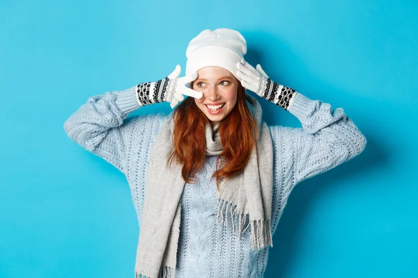 Концепция зимы и праздников. Симпатичная рыжая девушка-подросток в беании, перчатки и свитер с знаком мира, смотрит налево на пространство для копирования на синем фоне — стоковое фото