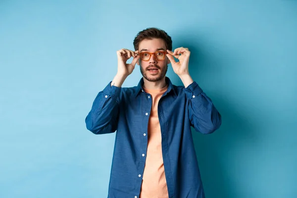 Imagem de belo homem caucasiano colocar em óculos e olhando para a câmera, tentando novos óculos na loja de óptica, fundo azul — Fotografia de Stock