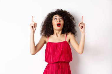 Gece makyajlı, kıvırcık saçlı, heyecanlı ve dik duran, reklam yapan, beyaz arka planda kırmızı elbiseli harika bir kız.