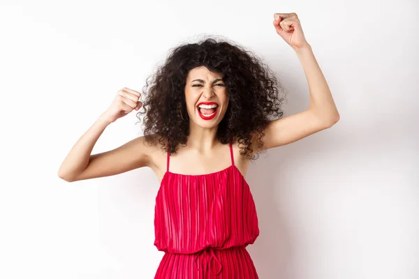 Vrolijke emotionele vrouw met krullend haar, rode jurk, handen omhoog en zingen, wroeten voor het team, schreeuwen willen winnen, staan op witte achtergrond — Stockfoto