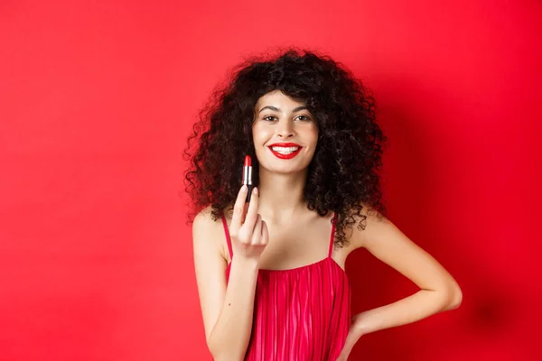 Modieuze vrouw met krullend haar, tonen rode lippenstift en glimlachen, aanbevelen cosmetische, staan op witte achtergrond — Stockfoto