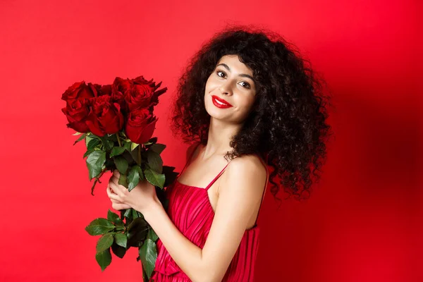 Felice bella donna in abito, tenendo fiori e sorridente romantico, in piedi sullo sfondo rosso — Foto Stock