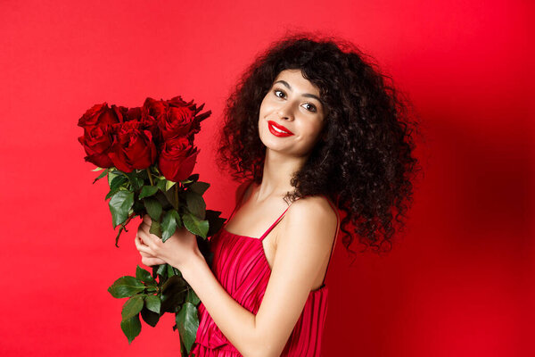 Happy beautiful woman in dress, holding flowers and smiling romantic, standing against red background
