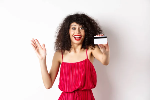 Winkelen. Verbazingwekkende krullende vrouw in rode jurk, tonen plastic credit card en schreeuw van opwinding, staan op witte achtergrond — Stockfoto
