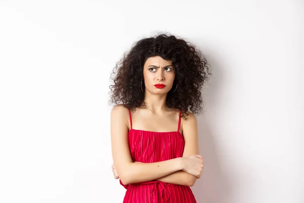 Suspeito e relutante mulher elegante, vestindo vestido vermelho e franzindo a testa, olhando para a esquerda com descrença, de pé sobre fundo branco — Fotografia de Stock