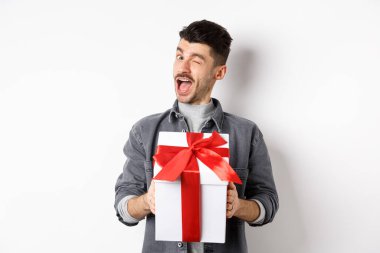 Sevgililer günü hediyesini tutan ve kameraya göz kırpan, aşığına romantik bir sürpriz yapan beyaz arka planda duran komik genç adam.