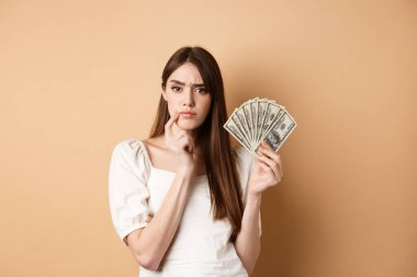 Ciddi ciddi para tutan ve düşünen, kameraya düşünceli görünen, kaşlarını çatan, dolar banknotlarıyla ne alacağına karar veren, bej arka plan