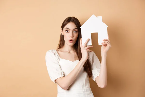 Real estate. Excited young woman showing house cutout and looking at camera, renting property, standing on beige background
