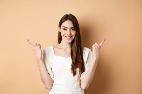 Mulher bonita em vestido apontando os dedos para o lado, mostrar duas maneiras, dando escolha e sorrindo, de pé sobre fundo bege — Fotografia de Stock