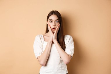 İnanılmaz. Etkilenmiş genç bir kadın nefes nefese kalmış ve haberlere hayran kalmış, kameraya bakıyor, huşu içinde yüzüne dokunuyor, bej arka planda duruyor.