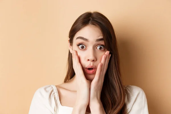 Face of surprised girl saying wow, touching cheeks and staring excited at camera, standing on beige background — Stock Photo, Image