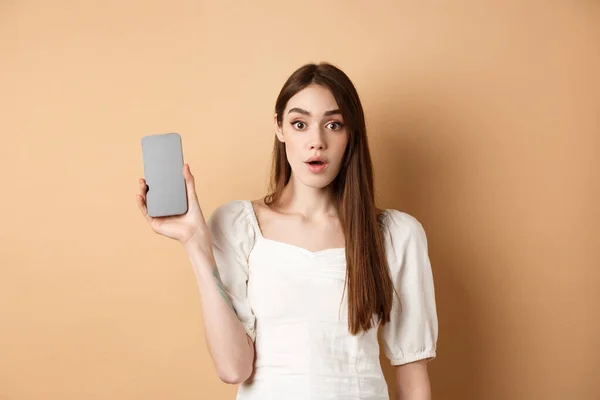 Garota bonita animado mostrando a tela do telefone celular vazio, olhar espantado com a câmera, de pé no fundo bege — Fotografia de Stock