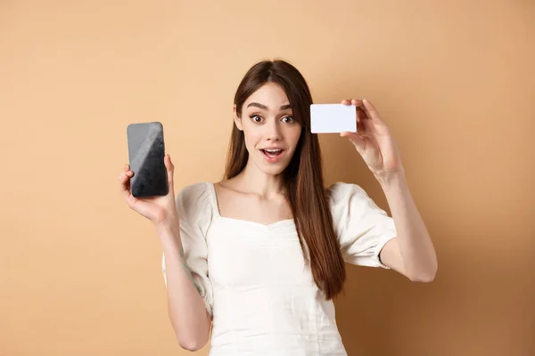 Garota entusiasmada mostrando cartão de crédito de plástico do banco e da tela do telefone celular vazio, demonstrar aplicativo de compras, de pé sobre fundo bege — Fotografia de Stock