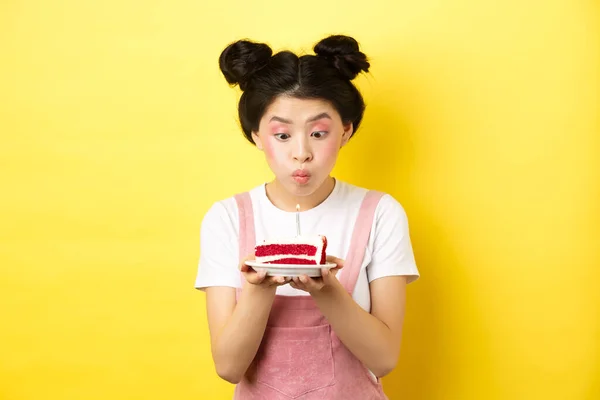 Праздники и праздники. Глупая азиатка с гламурным макияжем, загадывающая желание и задувающая свечу на торте ко дню рождения, стоящая на желтом фоне — стоковое фото