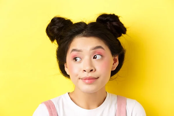 Close-up portret cute glamour azjatyckiej dziewczyny, jasny makijaż i fryzurę, patrząc na bok na logo z głupią twarzą, żółte tło — Zdjęcie stockowe