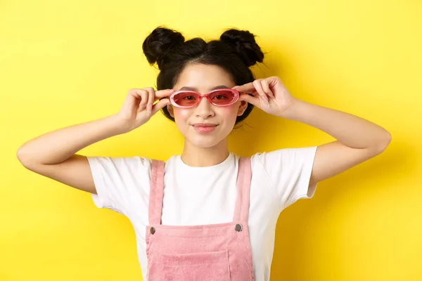 Hermosa chica asiática se puso gafas de sol con estilo y sonriendo, vistiendo traje de verano de moda, de pie sobre fondo amarillo — Foto de Stock