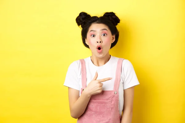 Vzrušená stylová asijská žena s půvabným make-upem, ukazující prstem doprava a lapající po dechu fascinována, řekněme, ohromen, žluté pozadí — Stock fotografie