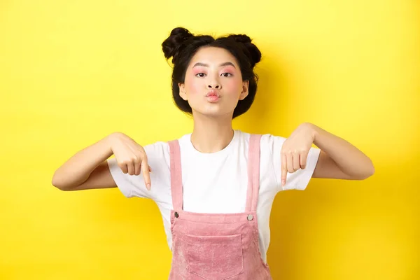 사시아 시안 십 대 소녀가 노란 배경에 매력적 인 화장과 여름 옷을 입고 서 있는, 아래에 있는 광고를 보고 손가락을 아래로 향하고 — 스톡 사진