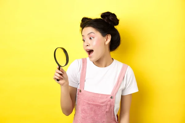 Opgewonden aziatische meisje vond interessant ding, kijkend door vergrootglas verbaasd, staande op gele zomer achtergrond — Stockfoto