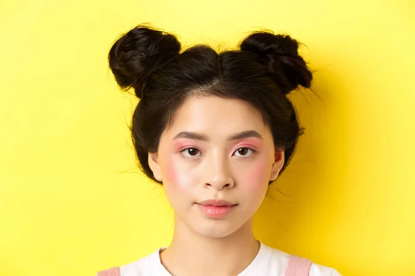 Retrato de cabeza de chica asiática con estilo con maquillaje brillante y bollos de pelo, mirando a la cámara, de pie sobre fondo amarillo —  Fotos de Stock