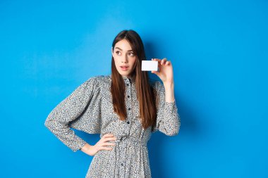 Alışverişe. Düşünceli bayan müşteri logoya bakıyor, plastik kredi kartını gösteriyor, mavi arka planda duruyor.