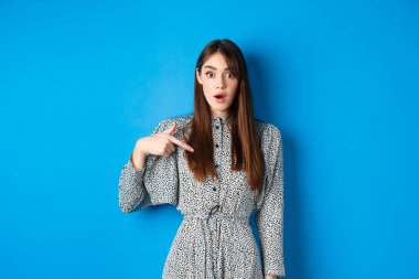 Şaşırmış genç bir kadın kendini inançsızlıkla işaret ediyor, seçiliyor ya da seçiliyor, mavi arka plana karşı elbise içinde duruyor.