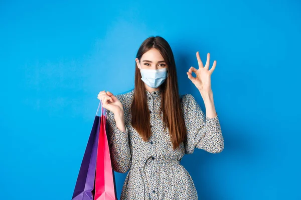 Covid-19, conceito de pandemia e estilo de vida. Mulher atraente usar máscara médica nas compras, mostrar ok e segurar sacos com compras, fundo azul — Fotografia de Stock