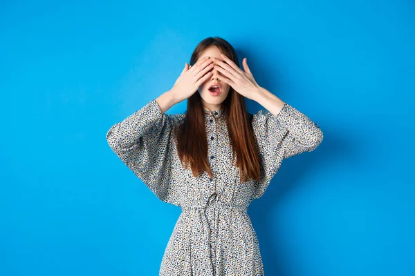 Izgalmas lány várja a meglepetést, miközben a szemét befedi a kezét, áll kíváncsi a kék háttér, ruha viselése — Stock Fotó