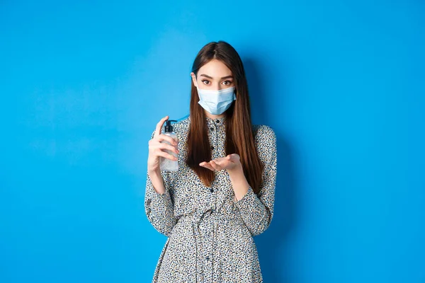 Covid-19, distanciamiento social y concepto de salud. Hermosa chica en máscara médica usando saniziter mano, sosteniendo la botella antiséptica, de pie sobre fondo azul — Foto de Stock