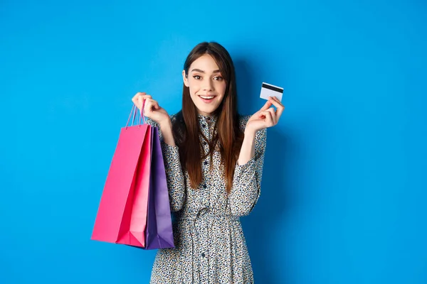 兴奋美丽的女模购物用塑料信用卡，拿着购物袋，高兴地看着相机，蓝色背景 — 图库照片
