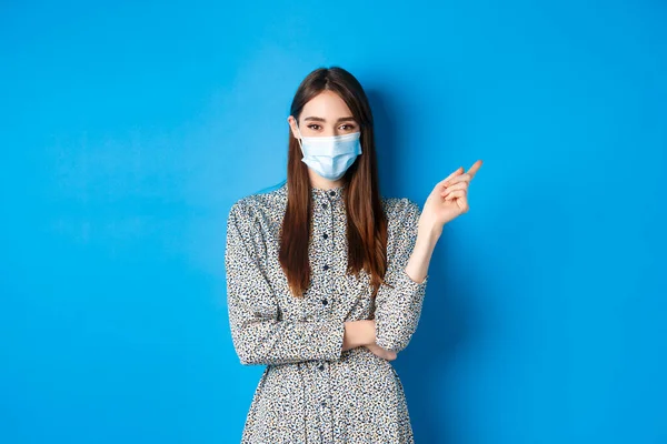 Covid-19, distanciamento social e conceito de saúde. Menina bonita em máscara médica mostrando caminho, apontando para a direita e sorrindo, publicidade em fundo azul — Fotografia de Stock